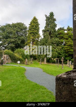 20. September 2026, Loweswater, Vereinigtes Königreich. Ein gewundener Kiesweg führt durch einen ruhigen Friedhof mit verwitterten Grabsteinen und üppigem grünem Gras Stockfoto