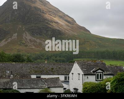 20. September 2026, Loweswater, Vereinigtes Königreich. Traditionelle Steinhäuser schmiegen sich an den Fuß eines rauen, mit Heidekraut bedeckten Hügels unter einer düsteren, übercasten Stockfoto
