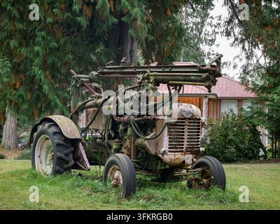 Alter verwitterter Traktor mit freiliegenden mechanischen Teilen steht auf Gras zwischen Bäumen, zeigt Rost, Alter und Jahrzehnte landwirtschaftlicher Nutzung. Stockfoto