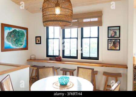 Helle Kauai-Essnische mit gewebter Rattan-Pendelleuchte über Holzbank, gerahmter Kartenkunst und drei Fenstern, die warmes Tageslicht hereinlassen, im Inneren Stockfoto
