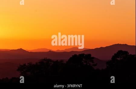 Entfernte Bergkämme und Täler Dunst füllen das Bild unter einem leuchtend orangefarbenen Himmel in Prescott Arizona, mit dunklen Baumsilhouetten in der Dämmerung über gestapelten Bergrücken Stockfoto