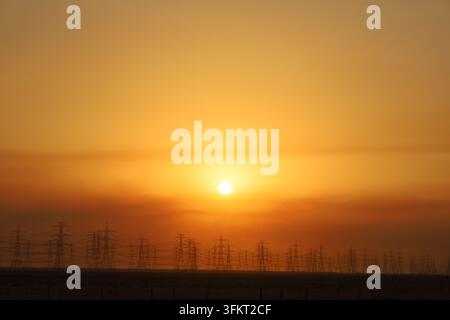 Silhouette von Strommasten und Hochspannungsübertragungsleitungen vor einem lebendigen goldenen Sonnenuntergangshimmel, ideal für Energie, Infrastruktur und Te Stockfoto