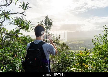 Wanderer von hinten gesehen zeigt Smartphone in Richtung des fernen grünen Tals und der Berge durch üppiges Dschungelfoliage auf Kauai Hawaii unter wolkenbeschienener Sonne in der Ferne Stockfoto