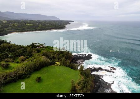 Oberblick auf die Landschaft von Kauai mit einem üppigen grünen Fairway, das zu einer dunklen vulkanischen Küste führt, wo die Pazifikwellen hereinrollen, mit felsigem schwarzem Lava und Cloß Stockfoto