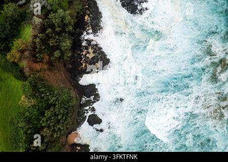 Vogelperspektive entlang Kauai zeigt türkisfarbenen Surf, der an dunklen Lavaklippen explodiert, üppige grüne Hügel und kleine Sandbuchten unten bei hellem Tageslicht coa Stockfoto