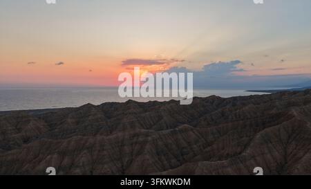 Luftaufnahme des Issyk-Kul-Sees bei Sonnenuntergang mit schroffem Berggelände im Vordergrund und einem dramatischen farbenfrohen Himmel in der Region Issyk-Kul, Kirgisistan. Stockfoto