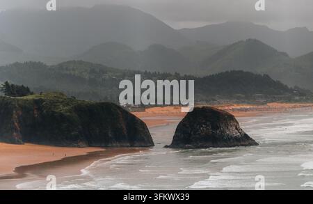 Luftbild von Haystack Rock an der sandigen Küste von Cannon Beach mit rollenden Wellen, üppigen grünen Küstenbergen und einem bewölkten Himmel in Cannon Beach Stockfoto