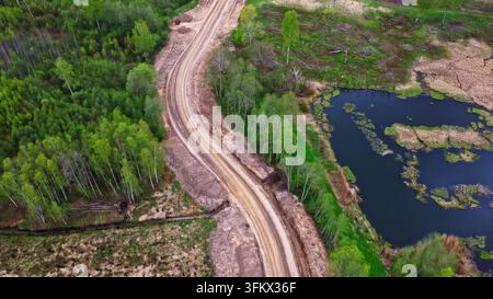 Luftaufnahme einer Schotterzugangsstraße, die sich durch einen üppigen grünen Wald neben einem dunklen Teich in Lettland windet. Stockfoto