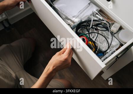 Hochwinklige Ansicht von Händen, die eine überfüllte weiße Schublade öffnen. Unordentliche Aufbewahrung mit verwirrten Kabeln, Ladegeräten und alter Elektronik. Konzept der Heimorganisation Stockfoto