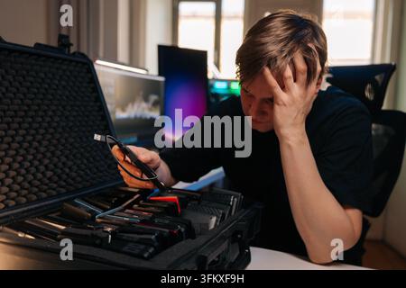 Besorgter junger Mann sitzt am Schreibtisch mit technischem Problem. Frustrierter Profi hält den Kopf und schaut auf viele externe Festplatten im Schutzkoffer Stockfoto