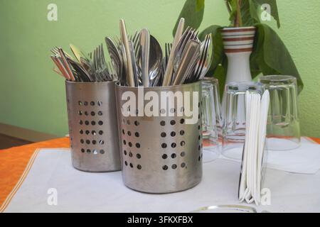 Edelstahlbesteck in perforierten Haltern neben Trinkgläsern und weißen Servietten auf einem Tisch. Stockfoto