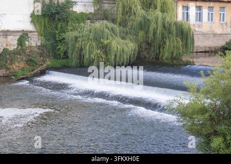 Flusswasser stürzt über ein stufenartiges Wehr unter herabhängenden Weidenzweigen neben alten Gebäuden. Stockfoto