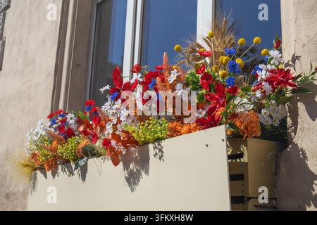 Bunte künstliche Blumen und getrockneter Weizen arrangiert in einer dekorativen Blumenkiste an einer sonnenbeschienenen Fassade. Stockfoto