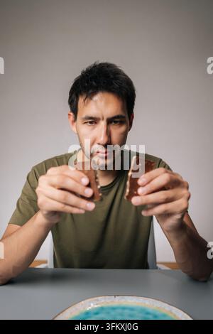 Vertikales Porträt eines nachdenklichen Mannes, der an einem Tisch sitzt und eine Schokoladenriege in zwei Stücke bricht, während er darüber nachdenkt, welches Stück er zuerst genießen soll Stockfoto