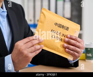 Nahaufnahme der Hände einer Geschäftsfrau, die ein Paket mit der Aufschrift „Made in China“ in chinesischen Schriftzeichen öffnet, während sie an einem Schreibtisch in ihrem Zuhause sitzt. Stockfoto