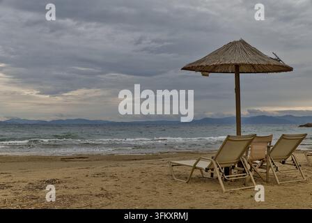 Leere Strandliegen unter einem verwitterten Strohsonnenschirm am Marikes Beach, Rafina, Ostattika, Griechenland, unter einem bewölkten Himmel. Stockfoto