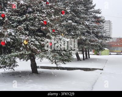 Eine Reihe von schneebedeckten immergrünen Bäumen, die mit roten und goldenen Weihnachtskugeln geschmückt sind, steht in einem städtischen Park während starken Schneefalls. Die Äste sind Stockfoto