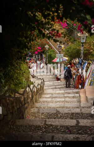 Taormina, Italien - 26. Juni 2026: Blick hinunter auf steinerne Treppen, die zum Strand Isola Bella in Taormina, Sizilien, führen, eingerahmt von blühenden Bougainvillea, Stände des lokalen Marktes, die den mediterranen Sommerurlaub und den italienischen Küstentourismus veranschaulichen Stockfoto