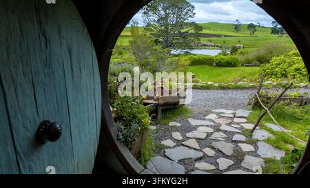 Blick durch die runde Tür eines Hobbit-Hochs, der über sanfte grüne Hügel auf dem Hobbiton-Filmset in Matamata, Waikato, Neuseeland hinausgeht Stockfoto