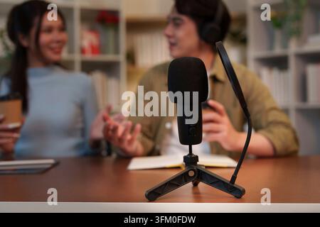 Nahaufnahme eines Desktop-Mikrofons mit Popfilter, mit einem verschwommenen asiatischen Mann und einer asiatischen Frau, die zusammen eine Podcast-Show moderieren, im Hintergrund Stockfoto