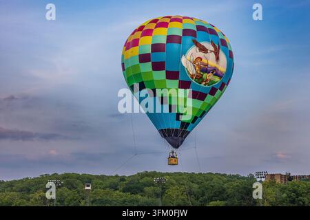 Ein farbenfroher Heißluftballon, geschmückt mit einer Darstellung eines Bogenschützen und fliegender Adler, steigt über eine baumgesäumte Landschaft auf. Stockfoto