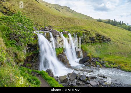 Der Gluggafoss-Wasserfall stürzt über moosige Felsen in einem lebendigen grünen Tal in Island, wobei kristallklares Wasser in einen felsigen Bach fließt. Stockfoto