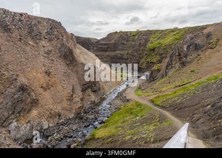 Ein gewundener Fluss fließt durch eine dramatische vulkanische Schlucht mit felsigen Klippen und grüner Vegetation in der Nähe von Husafell in Island an einem bewölkten Tag. Stockfoto