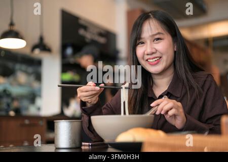 Eine horizontale Aufnahme einer jungen asiatischen Frau, die direkt in die Kamera schaut und ein helles, freundliches Lächeln zeigt. Sie ist bereit zu essen und hält ein Paar Essstäbchen Stockfoto
