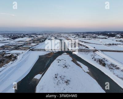 Eine schneebedeckte Landschaft mit einem Fluss, der durch sie fließt. Der Himmel ist eine Mischung aus Blau und Orange, was eine ruhige und friedliche Atmosphäre schafft Stockfoto