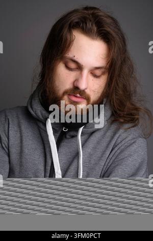 Studienporträt eines müden kaukasischen Mannes mit Bart und langen Haaren, der während eines grauen Hoodies die Augen schließt. Ideal für Themen wie Schlaf, Müdigkeit, Burnout Stockfoto