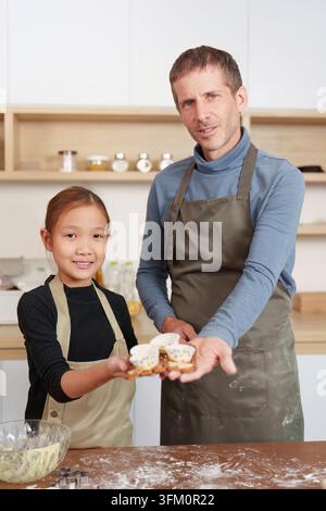 Ein Porträt eines lächelnden Vaters und einer Tochter, die Schürzen tragen und farbenfrohe Backförmchen in einer hellen modernen Küche darbieten Stockfoto