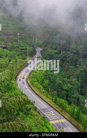 Luftaufnahme einer sich windenden zweispurigen Autobahn, die sich durch dichten tropischen Regenwald auf einem nebligen Berghang schneidet, mit sichtbaren Fahrzeugen auf der Straße in Mal Stockfoto