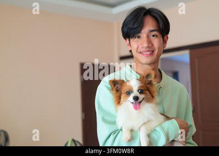 Ein horizontales Lifestyle-Porträt eines fröhlichen jungen asiatischen Mannes aus der Isan-Region Thailands, der drinnen steht. Sowohl der Mann als auch sein flauschiges Haustier Papil Stockfoto