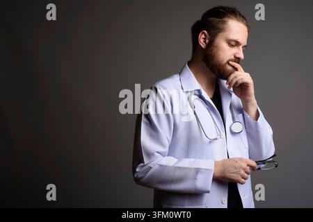 Studio-Porträt eines bärtigen jungen männlichen Arztes in einem weißen Labormantel mit Stethoskop, der seine Brille hält und nachdenklich nach unten blickt. Professionell Stockfoto