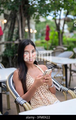 Eine vertikale Mittelaufnahme fängt eine schöne thailändische Frau ein, die in ein Café im Freien schaut und ihr Smartphone benutzt. Sie trägt ein stilvolles Polka Stockfoto