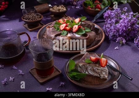 Schokoladen-Crêpe-Kuchen mit frischen Erdbeeren und Minze, begleitet von Kaffee und blühenden violetten Blumen, ein köstliches Dessert oder Frühstück s Stockfoto