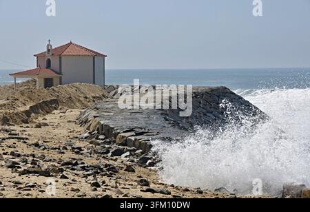 Kleine Kapelle, die auf einem sandigen Strand steht und durch einen Steinpier vor den brechenden Meereswellen geschützt ist Stockfoto