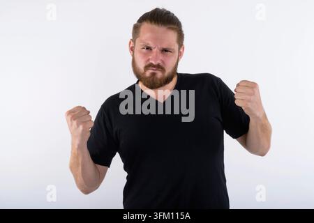 Studio-Porträt eines bärtigen jungen kaukasischen Mannes mit einem Haarbutt, der ein schwarzes T-Shirt trägt. Er schüttelt seine Fäuste mit einem wütenden, aggressiven Expre Stockfoto