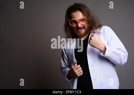 Studio-Porträt eines begeisterten bärtigen jungen Mannes mit langen Haaren, einem weißen Labormantel und einer übergroßen orangen Brille, der mit geballter Faust posiert Stockfoto