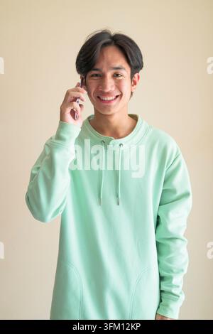 Ein Studio-Porträt eines fröhlichen jungen asiatischen Mannes aus der Isan-Region in Thailand, der ein Mobiltelefon an sein Ohr hält und breit lächelt Er sieht ernst aus Stockfoto