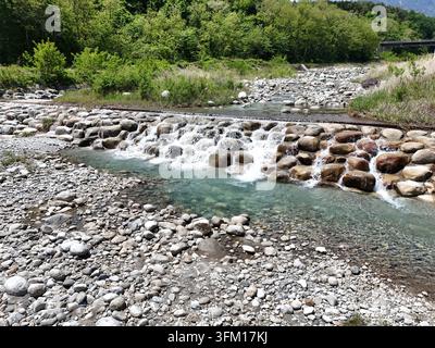 Der klare Strom fließt über glatte Steine. Die Vegetation am Flussufer schwankt sanft im Wind. Flaches Wasser zeigt Kieselsteine unter der Oberfläche. Das Sonnenlicht leuchtet Stockfoto