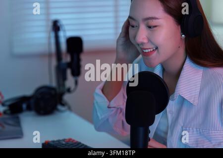 Ein Nahaufnahmeporträt einer lächelnden jungen asiatischen Frau, die Kopfhörer trägt und in ihr Heimstudio spricht Stockfoto