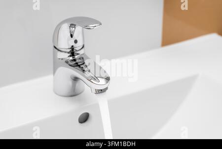 Wasser fließt aus einem chromfarbenen Badezimmertaph aus einem weißen Keramikwaschbecken in einem modernen Badezimmer Stockfoto