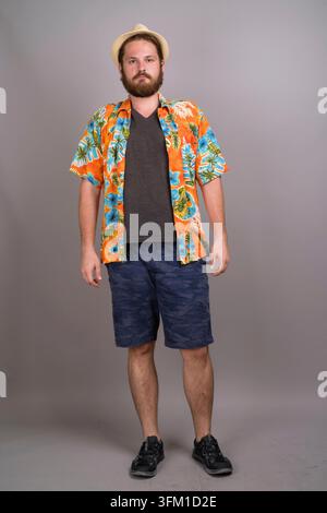 Vollständiges Studiofoto eines jungen kaukasischen Mannes mit Bart, der ein lebhaftes orangefarbenes Aloha-Hemd mit Blumenmuster, blaue Shorts und einen Trilby-Hut trägt. Perfekt für den Sommer Stockfoto