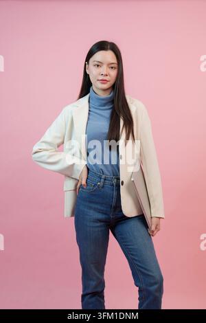 Mittlere Porträt einer professionellen asiatischen Frau in einem weißen Blazer und blauem Rollkragenpullover, die einen Laptop hält und selbstbewusst vor einem einfarbigen Rosa posiert Stockfoto