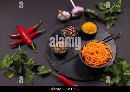 Koreanischer Karottensalat in einer Schüssel mit Paprika, umgeben von frischer Petersilie, Knoblauchzwiebeln, roten Chilischoten und kleinen Schüsseln mit bunten Gewürzen auf dunklem Hintergrund Stockfoto
