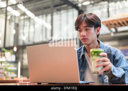 Horizontales, flaches Porträt eines ernsthaft fokussierten jungen südostasiatischen Thai-Isan-Mannes in einer Jeansjacke, der während der Arbeit ein Glas Matcha in der Hand hält Stockfoto