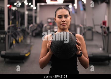 Eine Mittelaufnahme einer bestimmten jungen asiatischen Thai-Isan-Frau, die einen strukturierten schwarzen Medizinball mit beiden Händen vor ihrer Brust hält. Erfasst in einem Mod Stockfoto