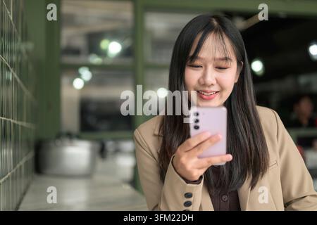 Eine horizontale Medium-Klappaufnahme, die eine junge asiatische Frau zeigt, die mit einem fröhlichen Lächeln auf ein rosa Smartphone in ihrer Hand schaut. Sie trägt ein Stockfoto