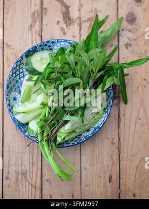 Eine Top-Down-Ansicht von frischen thailändischen aromatischen Kräutern, darunter süßer Basilikum, Morgenröschen und geschnittene Gurken, serviert in einer blauen Keramikschüssel auf rustikalem Holz Stockfoto